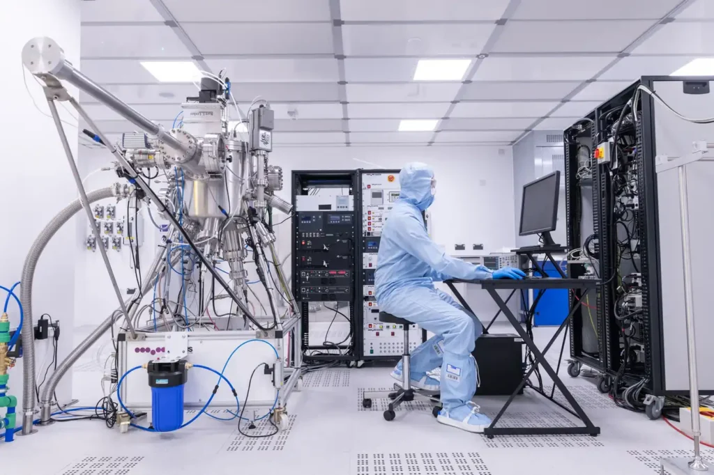 Researcher in cleanroom clothing working at a computer beside specialist scientific equipment and server racks.