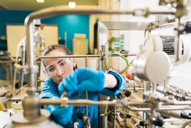 Researcher wearing safety glasses and blue gloves adjusting stainless steel process engineering equipment in a laboratory.
