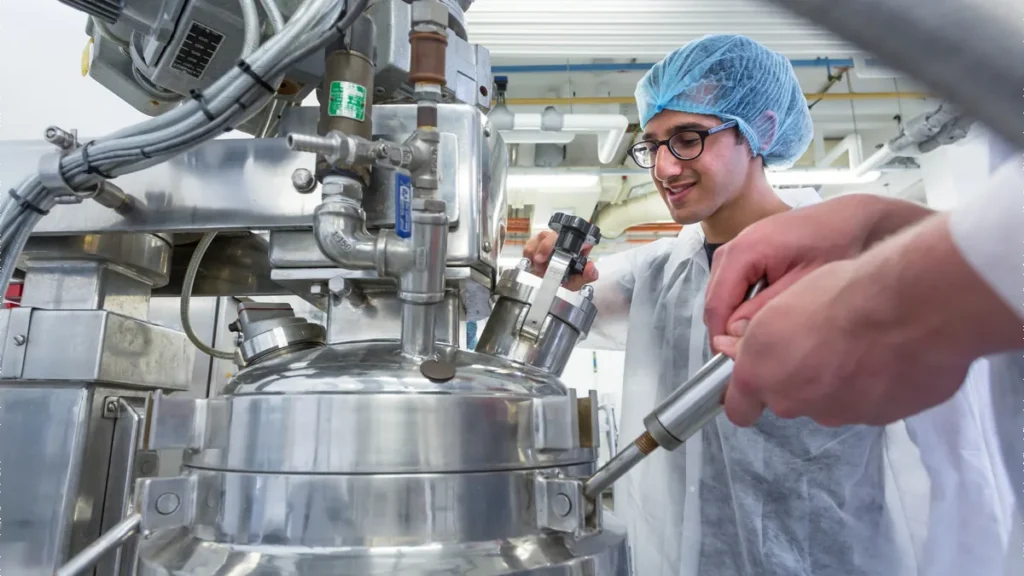 Food production specialist wearing a hair net inspecting stainless steel processing equipment in a laboratory setting.