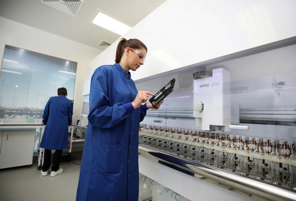 Researcher wearing safety glasses and a blue lab coat using a handheld device beside automated laboratory equipment.