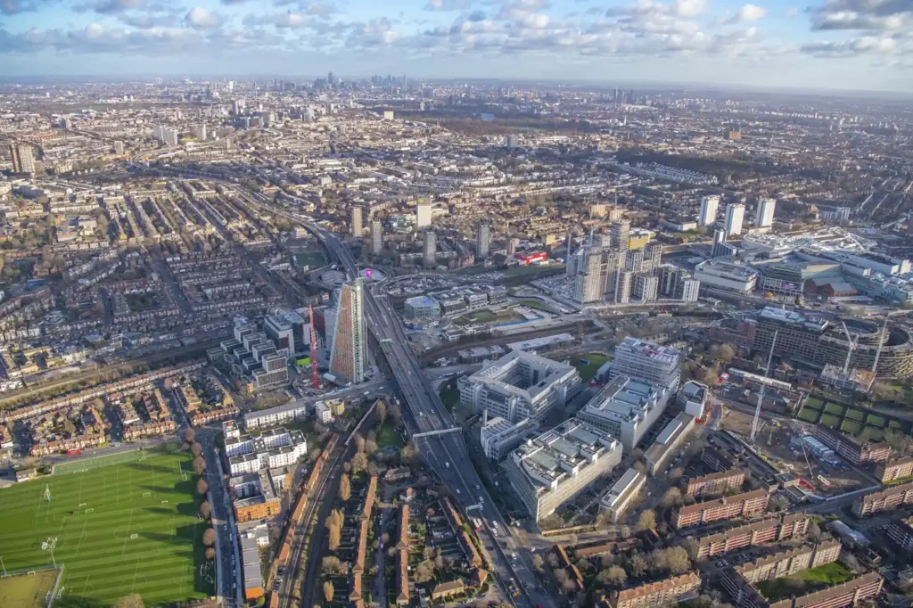 Aerial view across a dense urban London district with roads, housing, sports fields and high-rise buildings.